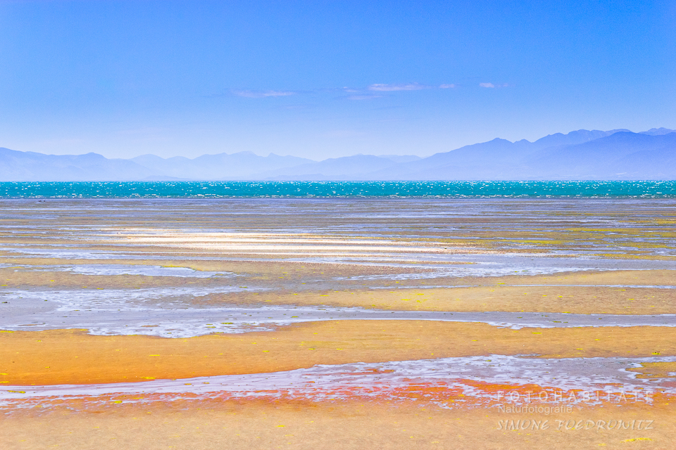 buntfarbener Strand bei Ebbe vor türkisem Meer und Bergen